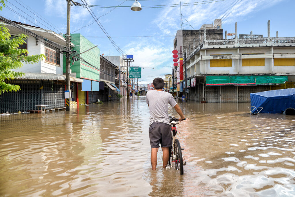 A man stands ankle-deep in floodwater on a street, holding a bicycle. The street is lined with closed shops and buildings, some with metal shutters down. The road is heavily flooded, with water reflecting the buildings and sky. A blue sky with scattered clouds is visible above, and the atmosphere appears calm despite the flooding.