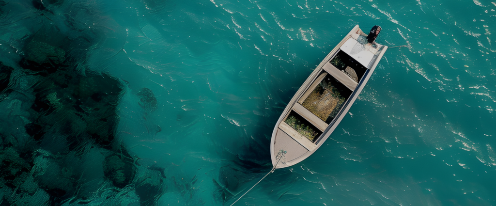 Aerial view of a small motorboat floating on clear turquoise water near a darker patch of reef or rocks. One person is seated at the back of the boat, and the scene highlights the contrast between shallow and deeper water areas.