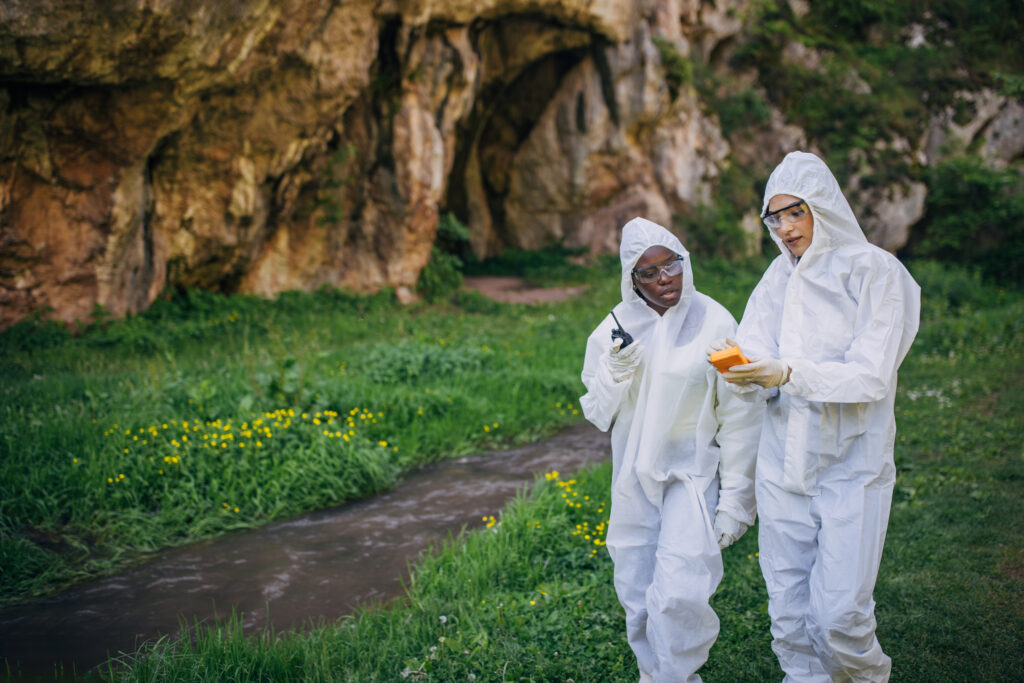 Two people wearing white protective suits and gloves walk through a grassy area near a cave entrance. One person holds an orange object while the other listens attentively. The scene suggests a scientific or environmental field investigation.