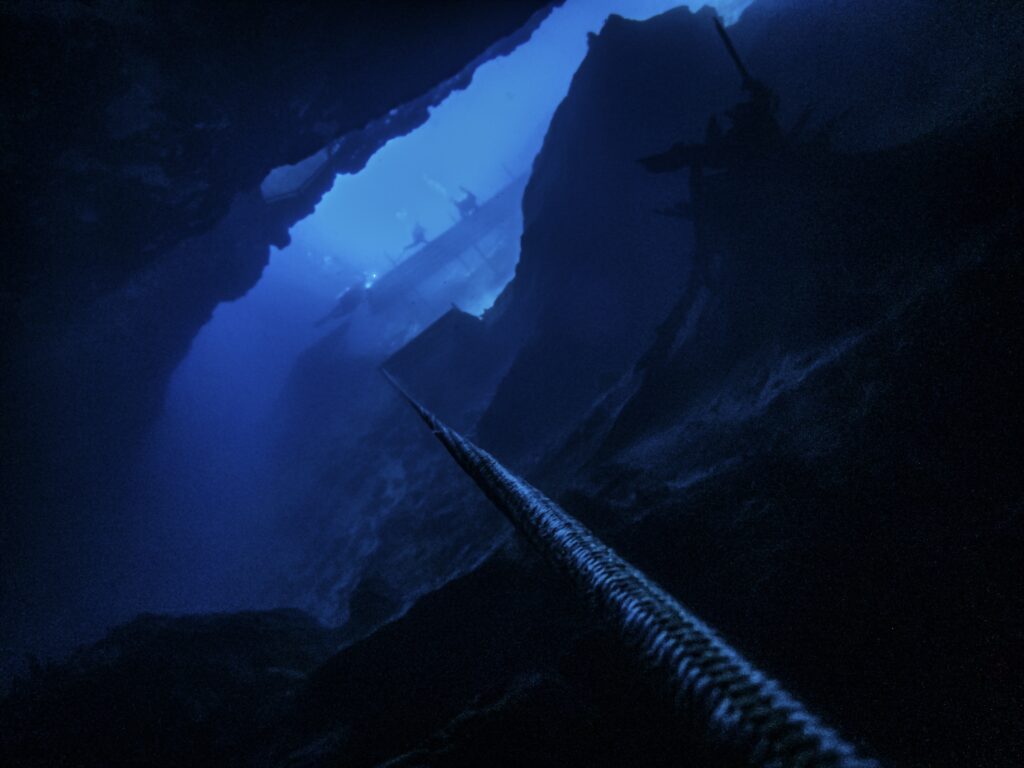 A dark, underwater scene showing a large cavern or crevice with a rope extending downwards into the depths.