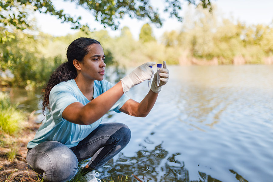A woman wearing gloves kneels beside a body of water, holding up a test tube with liquid inside, appearing to be taking a watersample