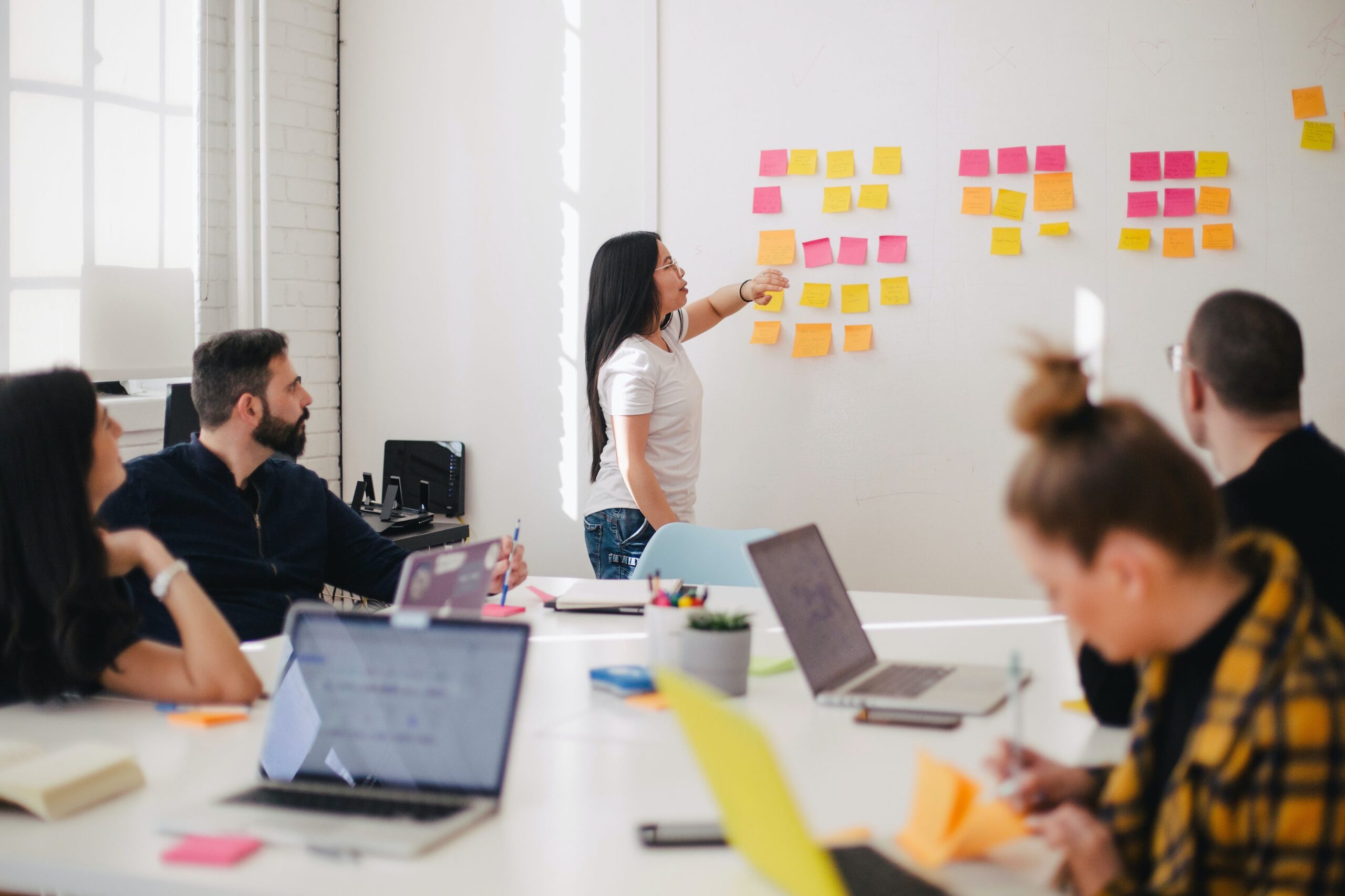 A group of five people in a modern office meeting room are gathered around a table with laptops and notepads. One woman stands at the front, pointing to a wall covered in colorful sticky notes, leading a brainstorming or planning session. The others are seated, listening, taking notes, or working on laptops. Let me know if you need a shorter version or one tailored to a specific context (e.g., workshop, tech team, classroom).