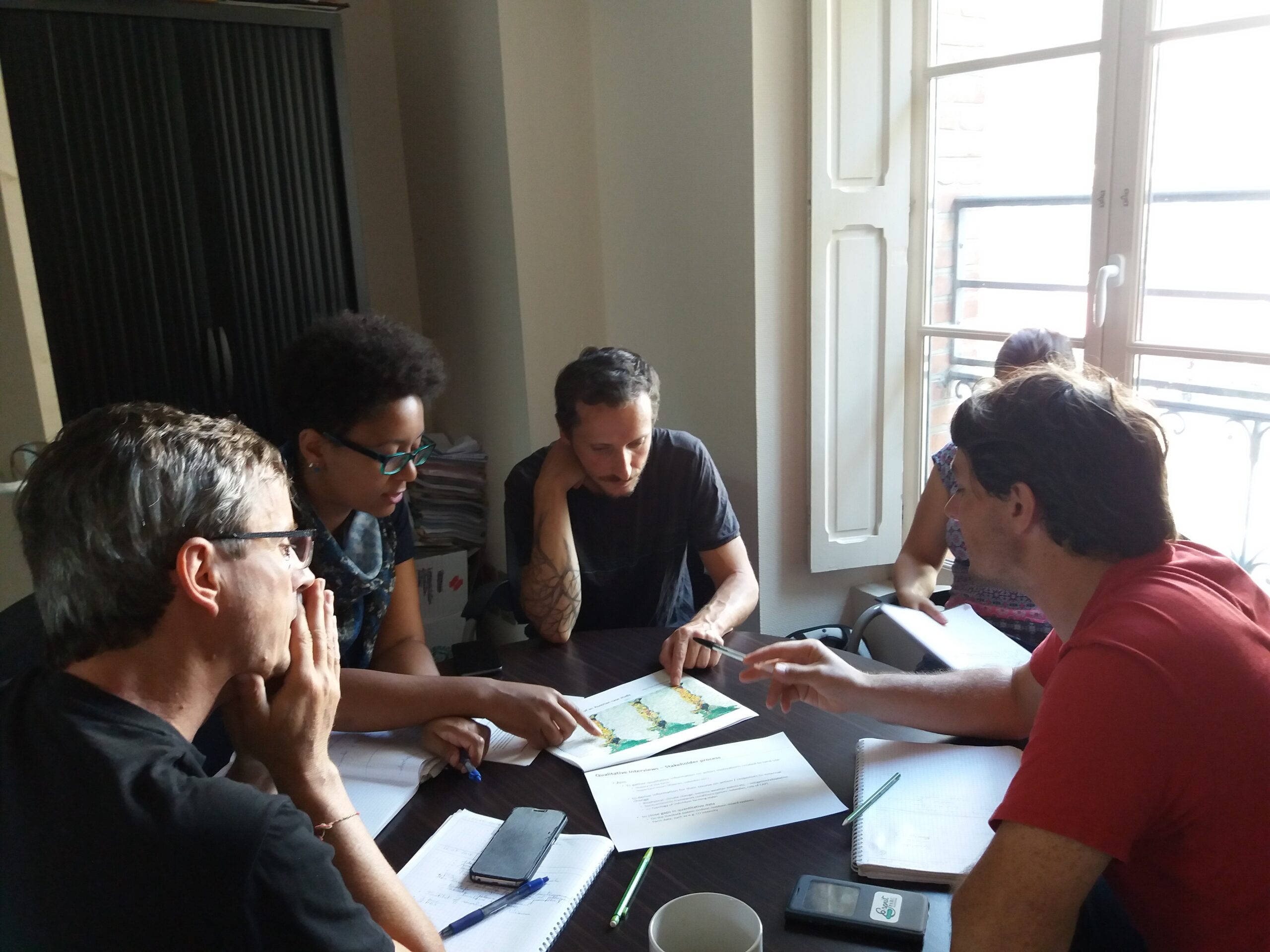 Five people of the P3 - People, Pollution and Pathogens project sit around a table in a well-lit room, engaged in a focused discussion. Papers, notebooks, and laptops are spread out on the table, and one person gestures toward a document as others listen or look on attentively. Natural light enters through a large window in the background.