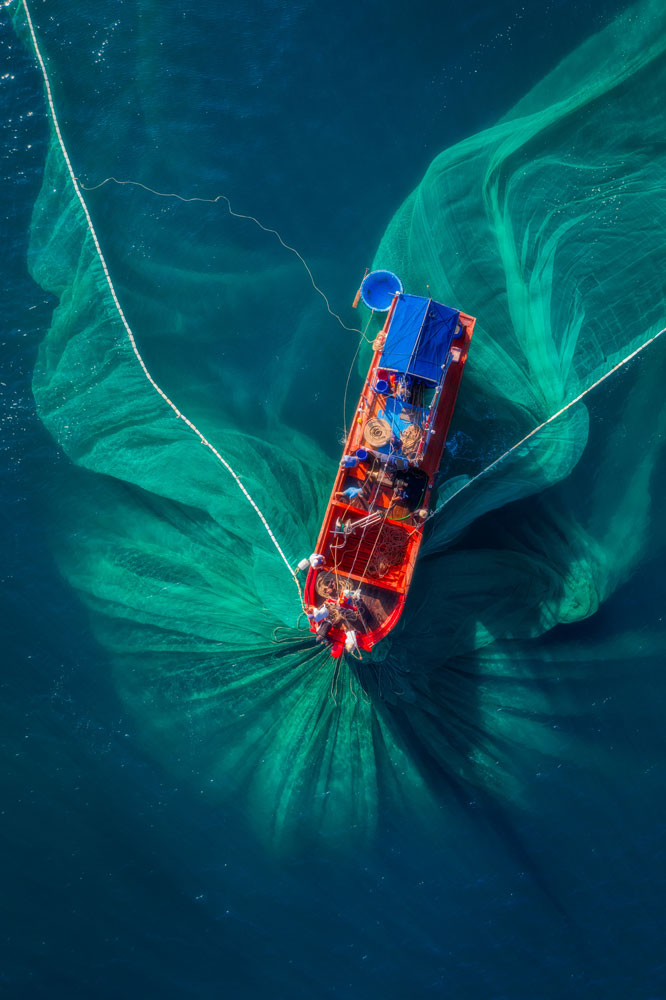 Aerial view of a brightly colored fishing boat casting wide green nets into deep blue water. The nets create sweeping patterns around the boat as several crew members work on deck. The scene highlights traditional fishing activity from above.