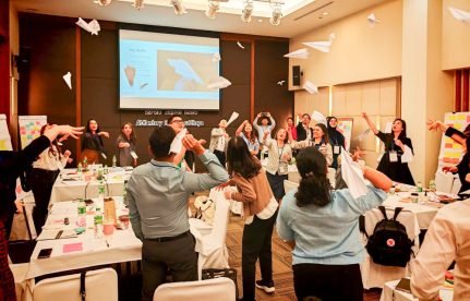A lively conference room filled with people standing and tossing paper airplanes into the air. Tables are covered with workshop materials and name tags. A large screen at the front displays a slide with an image of a paper airplane and a speaker’s photo. The atmosphere appears interactive and energetic.